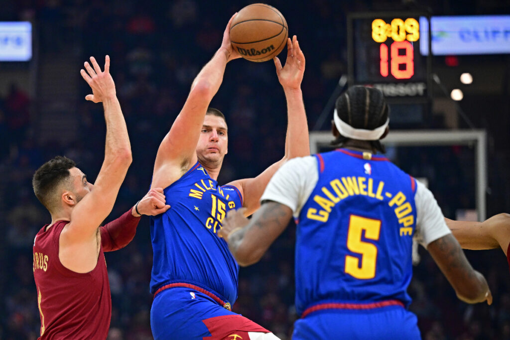 Denver Nuggets center Nikola Jokic, center, attempts to pass to Kentavious Caldwell-Pope while being defended by Cleveland Cavaliers guard Max Strus in the first half of an NBA basketball game, Sunday, Nov. 19, 2023, in Cleveland. (AP Photo/David Dermer)