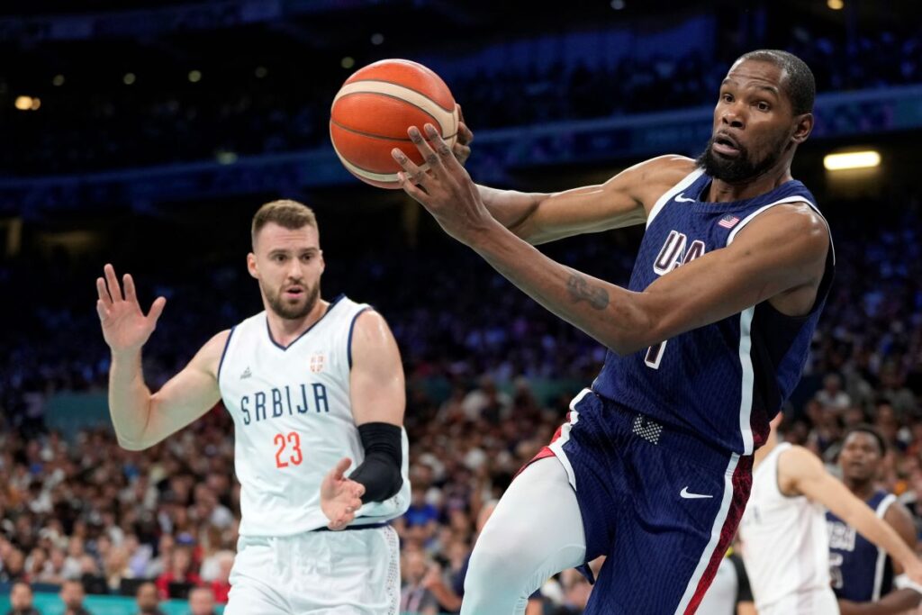 Kevin Durant, right, of the United States, passes the ball as he heads out of bounds while under pressure from Marko Guduric, of Serbia, in a men's basketball game at the 2024 Summer Olympics, Sunday, July 28, 2024, in Villeneuve-d'Ascq, France. (AP Photo/Michael Conroy)