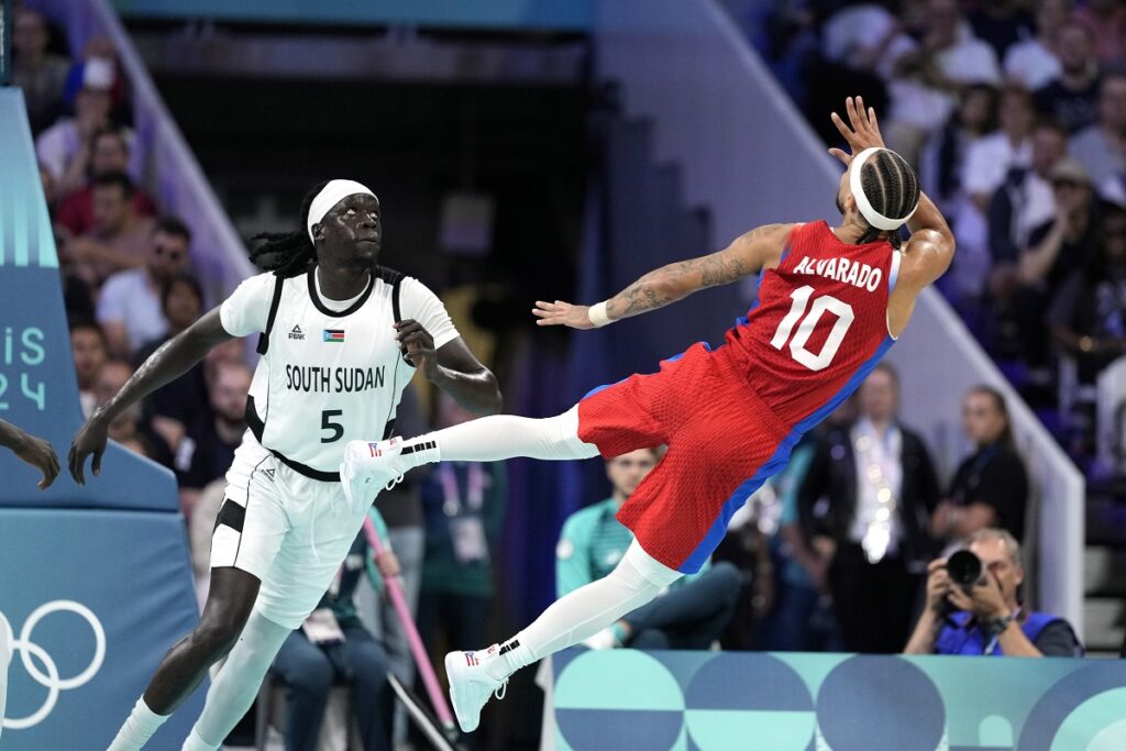 Jose Alvarado, right, of Puerto Rico, flies through the are after shooting as Nuni Omot, of South Sudan, defends in a men's basketball game at the 2024 Summer Olympics, Sunday, July 28, 2024, in Villeneuve-d'Ascq, France. (AP Photo/Michael Conroy)